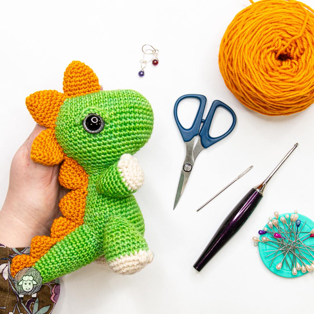 green, orange, and white crocheted amigurumi dinosaur wtih craft supplies in background